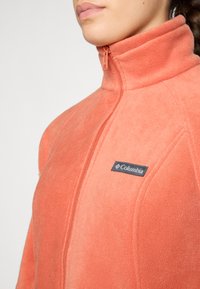 Veste en polaire de couleur corail avec un col haut, fermeture éclair sur toute la longueur à l'avant et un patch logo Columbia noir sur la poitrine gauche. Texture douce.