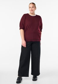 Burgunderroter gestrickter Pullover mit kurzen Ärmeln und rundem Halsausschnitt. Kombiniert mit weiten schwarzen Hosen und schwarzen Loafers mit Nietenakzenten.