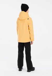 Veste imperméable jaune avec capuche et logo au dos, associée à un pantalon et des chaussures noirs, sur un fond blanc uni.