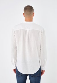Camicia bianca in lino con maniche lunghe, orlo rotondo e dettaglio sul retro. Presenta bottoni sui polsini e un tessuto leggero e con texture.