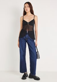 Melna caurspīdīga kamizole ar priekšpuses mezglu un apļveida akcentu, kopā ar tumši ziliem augsta vidukļa džinsiem un melnām masīvām loaferiem.