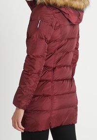 Abrigo acolchado color granate con capucha adornada con piel, diseño acolchado, bolsillos laterales con cremallera y corte a media longitud, que proporciona calidez y cobertura.