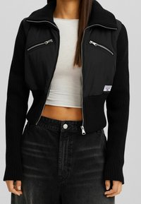 Chaqueta negra corta con parte superior acolchada, dos bolsillos en el pecho con cremallera, mangas acanaladas y una cremallera plateada. Se lleva sobre una camiseta clara.