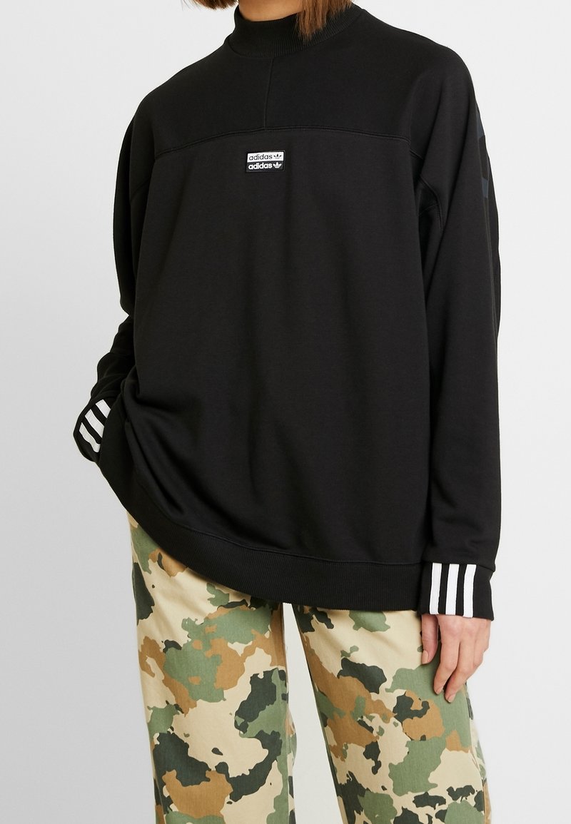 Černá mikina s vysokým límcem, bílý logo adidas na hrudníku, žebrované manžety s třemi bílými pruhy, doplněná o kalhoty s maskáčovým vzorem.
