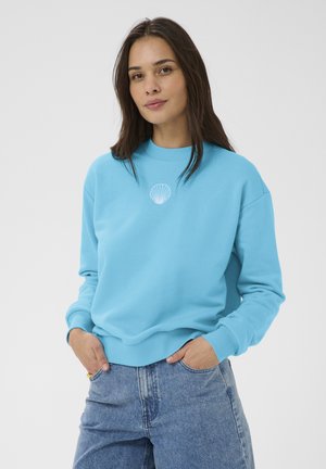 Kvinde med langt brunt hår iført en lyseblå sweatshirt med et hvidt skaldyrsmotiv, hænderne i lommerne og blå jeans mod en ensfarvet baggrund.