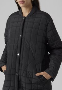 Chaqueta de plumas acolchada negra con cuello alto, botones de presión en el frente y bolsillos laterales. La tela parece suave con un sutil brillo.