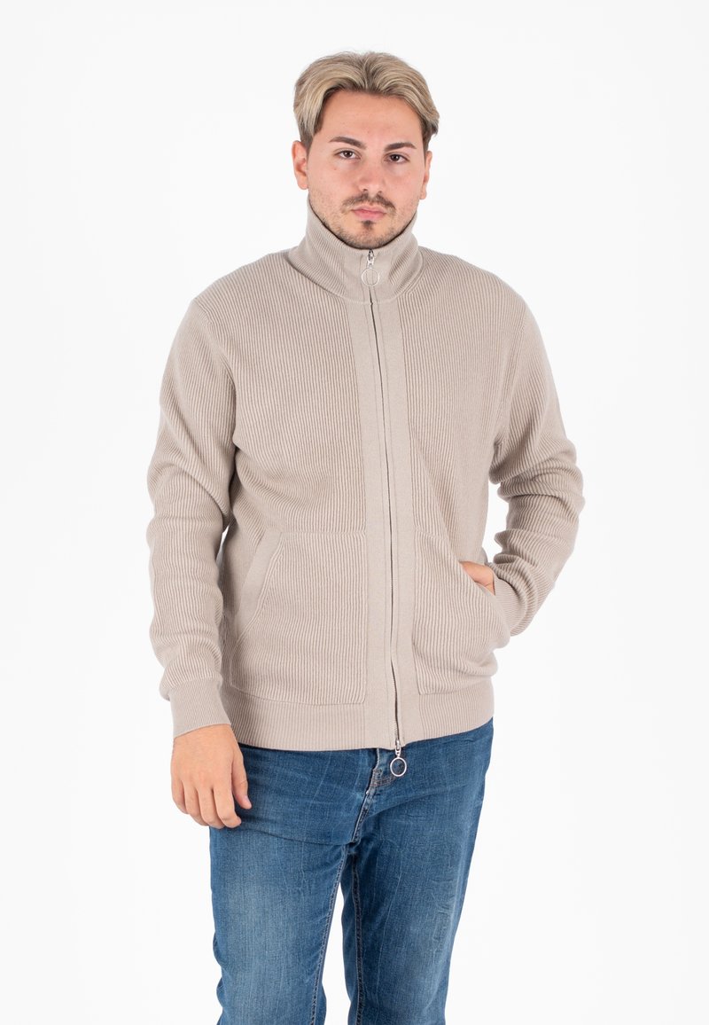 Beige geribde ritsjack met hoge kraag, twee voorzakken en een zilveren ring trekrits, gecombineerd met blauwe jeans.