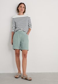 Pruhovaná mikina s dlouhými rukávy v bílé a námořnické barvě, spojená se světle zelenými lněnými šortkami. Šedé sandály s křížovým designem popruhů. Jednoduché pozadí.