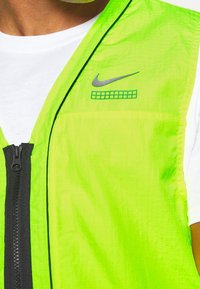Ljus neongrön Nike-väst med dragkedja fram, svart kant och reflektiva logodetaljer. Lätt, texturerad tyg med en minimalistisk design.