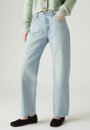 Persoon die lichtblauwe jeans met rechte pijpen, een groene gebreide trui en zwarte balletschoenen draagt, met één hand in de zak, neutrale achtergrond.