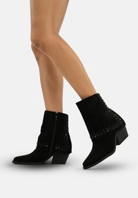 Botas de tobillo de ante negro con tacón grueso, que presentan cremalleras laterales y acentos de tachuelas a lo largo de la parte superior. Textura suave y diseño de punta afilada.