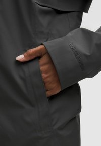 Pilkas vandeniui atsparus švarkas su kišene su dangteliu, rodančia ranką, besistengiančią pasiekti kišenę. Rankovėse matomas subtilus Lululemon logotipas.