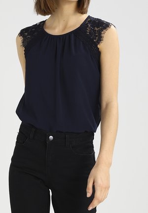 Vrouw draagt een mouwloos marineblauw blousje met kanten schouders, ingestopt in een zwarte jeans met hoge taille, staand tegen een effen achtergrond.