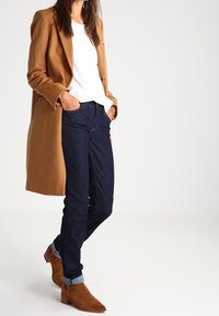 Cappotto di lana marrone, camicia bianca a maniche lunghe con trama, jeans in denim scuro, risvolti sulle maniche, e stivali alla caviglia in suede marrone con tacco basso.