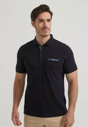 Homme portant un polo noir à manches courtes avec un pantalon beige, debout et regardant légèrement vers la droite sur un fond clair uni.