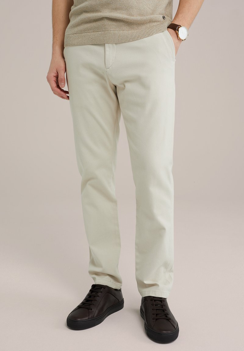Light beige trousers made of soft fabric, featuring a slim fit, with a smooth texture and side pockets, paired with dark brown sneakers.