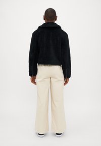 Schwarze, flauschige, kurz geschnittene Jacke mit Schalkragen, von hinten gezeigt. Kombiniert mit beigen weit geschnittenen Hosen und schwarzen Sneakers.