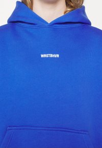 Personne portant un sweat à capuche bleu vif avec un texte blanc "WRSTBHVR" sur la poitrine et une poche avant visible.