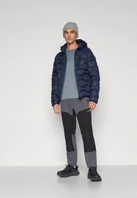 Vīrietis, kurš valkā pelēku cepuri, tumši zilu pūkainu jaku, gaiši zilu kreklu, pelēkas un melnas krāsas bikses un melnus sporta apavus, stāv pret vienkāršu baltu fonu.