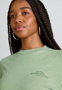 Ανοιχτό πράσινο μπλουζάκι φτιαγμένο από βαμβάκι, με στρογγυλή λαιμόκοψη και μικρό εκτυπωμένο λογότυπο σε σκουρότερη απόχρωση μπροστά.