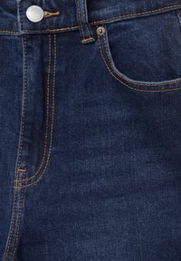 Primer plano de unos jeans de mezclilla azul oscuro que muestra el botón de metal, el bolsillo delantero, el lazo del cinturón y los detalles de costura en naranja.