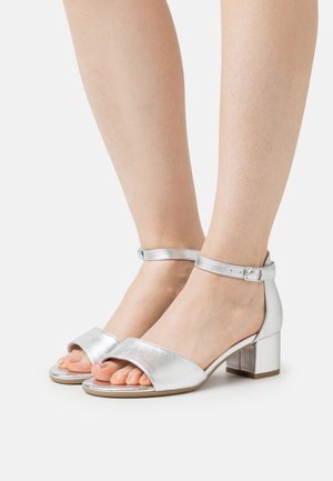 Sandales argentées à talons bas avec des brides de cheville, portées aux pieds, mettant en valeur un design ouvert à l'avant et des talons carrés, sur fond blanc uni.