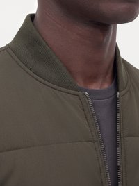 Detail osoby s krkom a čeľusťou, ktorá má na sebe tmavozelenú bundu na zips cez sivú košeľu na svetlom pozadí.