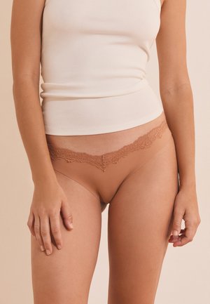Femme portant des sous-vêtements beige avec dentelle et un débardeur blanc côtelé, debout devant un fond neutre uni.