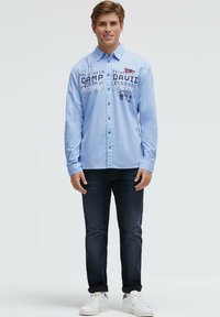 Hellblaues Langarmhemd mit Knopfleiste und bedruckten Grafiken. Kombiniert mit dunklen Jeans und weißen Turnschuhen. Einfaches, lässiges Design.