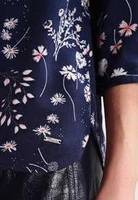 Tissu bleu marine avec des motifs floraux blancs et roses sur une manche de chemise au-dessus d’un pantalon noir texturé, montrant une petite étiquette métallique indiquant « Denim ».