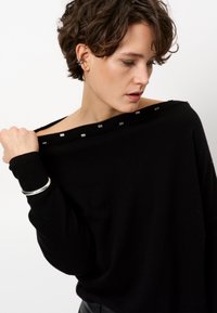 Schwarzer übergroßer Pullover mit weicher Textur, schulterfreier Gestaltung und silbernen quadratischen Akzenten am Ausschnitt.