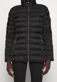 Svart, vadderad pufferjacka med dragkedja fram och dragkedjefickor, bärs av en person med mörk hud och svarta byxor.