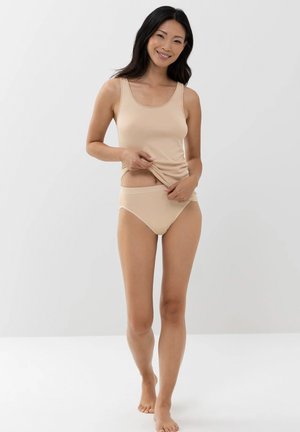 Lächelnde Frau in beigem Tanktop und Unterwäsche, die ihr Oberteil anpasst, barfuß vor einem einfarbigen hellgrauen Hintergrund stehend.