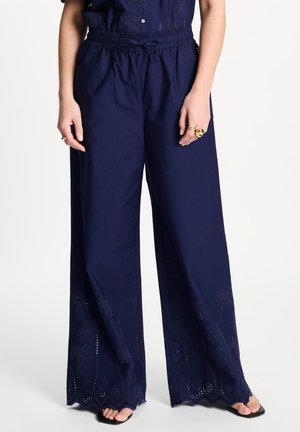 Vrouw draagt marineblauwe broek met brede pijpen, geborduurde zoom en elastische tailleband, gecombineerd met zwarte open-toe schoenen en gouden ringen aan de vingers.