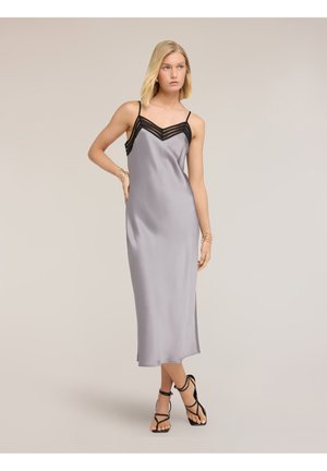 Frau trägt ein ärmelloses silbernes Satin-Midikleid mit schwarzem Spitzenbesatz und schwarzen Riemchenabsätzen, posiert vor einem schlichten Hintergrund.