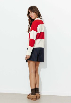 Pull rayé rouge et crème associé à un short bleu marine à fines rayures blanches. Porté avec des chaussures marron et des chaussettes foncées. Le mannequin est tourné de dos, mettant en valeur la tenue.