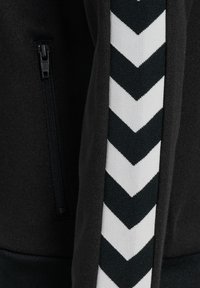 Giacca atletica nera con tasca con zip e pannello laterale con motivo a chevron bianco su un tessuto testurizzato.