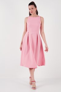 Sieviete, tērpusies bezroku rozā midi kleitā, ar daudzkrāsainām augstpapēžu sandales un ziedu formas auskariem, stāvot pret balto fonu.