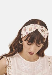 Diadema de tela con estampado floral en color crema, presenta un diseño de nudo torcido; complementada con unos pendientes de aro dorados y una blusa de encaje delicada.