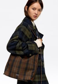 Tote torba v temno rjavi barvi s karirastim vzorcem, ki vključuje črne in rdeče črte. Narejena iz mehkega materiala, ima ohlapen obliko in usnjene dodatke.