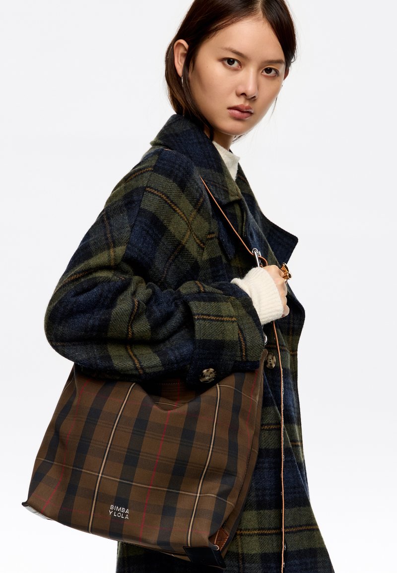 Tote torba v temno rjavi barvi s karirastim vzorcem, ki vključuje črne in rdeče črte. Narejena iz mehkega materiala, ima ohlapen obliko in usnjene dodatke.