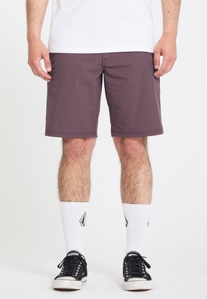 Hombre con pantalones cortos morados, calcetines blancos hasta el tobillo con un logo negro, zapatillas negras de corte bajo y camiseta blanca, de pie contra un fondo blanco.