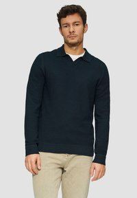 Marineblauw gebreid pullover met V-hals en kraag, voorzien van ribgebreide manchetten en zoom, gedragen over een witte blouse, gecombineerd met lichtbeige broek.