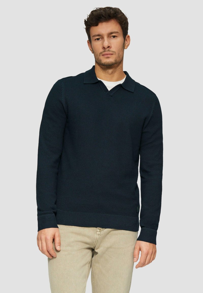 Marineblauer strukturierter Strickpullover mit V-Ausschnitt und Kragen, mit gerippten Bündchen und Saum, kombiniert mit einem weißen Hemd und hellbeigen Hosen.