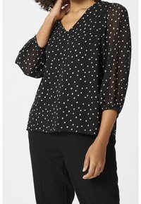 Blouse noire à pois blancs, avec un décolleté en V et des manches bouffantes transparentes. Le tissu semble léger avec une texture lisse.
