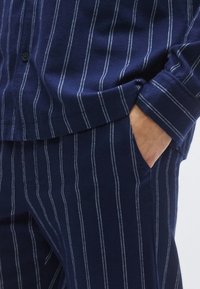 Granatowy zestaw piżamowy z miękkiego materiału z cienkimi białymi paskami w pionie. Koszula posiada zapięcie na guziki; spodnie mają boczne kieszenie.