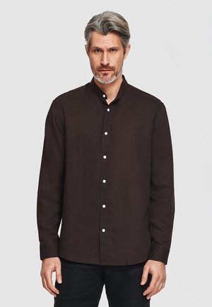 Mężczyzna z siwymi włosami i brodą, ubrany w zapinaną na guziki ciemnobrązową koszulę z długim rękawem, stojący na tle jednolitego jasnego tła.