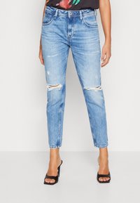 Jeans strappati azzurro chiaro con gamba affusolata, caratterizzati da tagli sfrangiati sulle ginocchia e una leggera décoloration. Abbinati a tacchi neri aperti in punta.