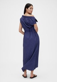 Abito lungo blu navy con spalle scoperte e pois bianchi, vita aderente e gonna fluente. Indossato con sandali neri. Tessuto dalla texture liscia.