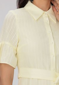 Robe à boutons couleur jaune clair, avec un col, des manches courtes bouffantes et des rayures verticales. Tissu doux avec une taille ceinturée.